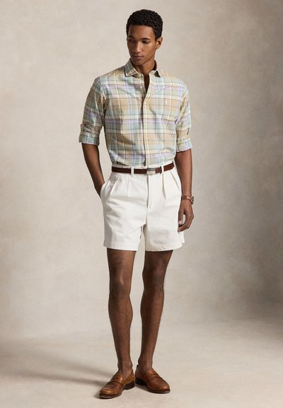 Homme portant une chemise à carreaux beige avec manches retroussées, short plissé blanc, ceinture marron et mocassins marron, debout sur un fond neutre.