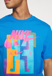 Blå t-shirt med stor färgglad Nike Air grafik i rosa, orange och blått, som bärs av en person med synlig hals och delvis ansikte.