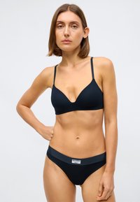 Jeune femme aux cheveux courts bruns portant un soutien-gorge noir et un bas noir assorti, se tenant contre un fond clair uni.