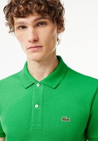 Green polo shirt made of textured fabric, featuring a collar and two buttons. Embroidered crocodile logo on the left chest.