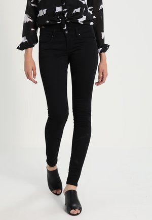 Vrouw draagt zwarte skinny jeans, zwarte open-toe mule hakken en een zwart-wit patroon blouse, staand tegen een witte achtergrond.