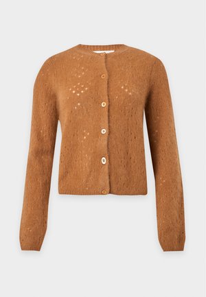Femme portant un cardigan orange boutonné, un jean large bleu, des talons noirs pointus, tenant un sac à main noir sur un fond uni.