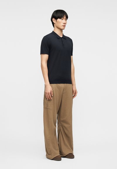 Homme debout portant un polo noir à manches courtes, un pantalon cargo ample couleur beige, et des chaussures marron, sur un fond gris uni.