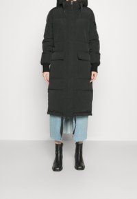 Svart puffjacka med huva, lång längd, har två framfickor, ribbade ärmslut och dragsnodd i nederkant. Kombination med en denimkjol och svarta stövlar.