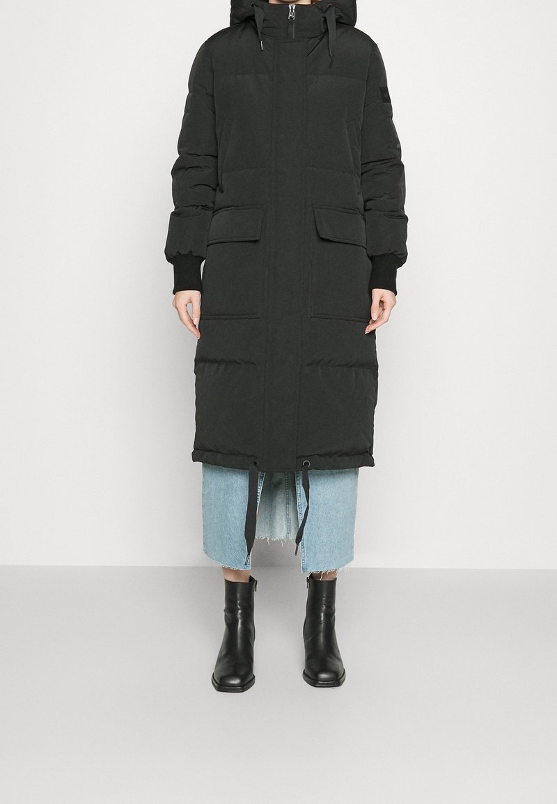 Svart puffjacka med huva, lång längd, har två framfickor, ribbade ärmslut och dragsnodd i nederkant. Kombination med en denimkjol och svarta stövlar.