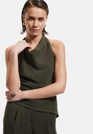 Olivgrünes Trägershirt mit drapierter Ausschnittlinie und weichem Stoff. Die Arme des Models sanft vor der Taille gekreuzt, was das elegante Design zur Geltung bringt.