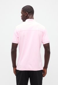 Άνδρας με πλάτη, φορώντας ένα ανοιχτό ροζ κοντομάνικο πουκάμισο πόλο και μαύρο παντελόνι, σε απλό λευκό φόντο.