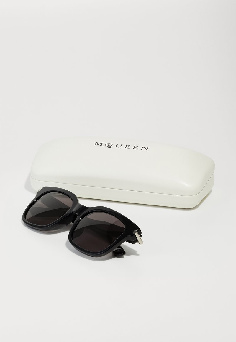 Lunettes de soleil noires carrées avec des verres foncés, présentant une finition mate élégante et des accents argentés, placées à côté d'un étui rigide blanc étiqueté "M QUEEN".