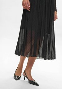 Rochie midi cu buline negre, dintr-un material transparent, cu un design stratificat, asortată cu sandale negre lucioase cu toc subțire și vârf ascuțit.