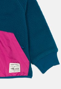Veste en polaire teal avec une poche rose. Présente des poignets côtelés et une étiquette tissée en bas. Texture douce avec une finition légèrement duveteuse.