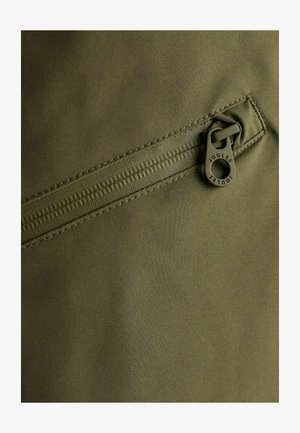 Tela verde con un bolsillo con cremallera que tiene un tirador redondo etiquetado "Joules". La textura es suave, con una costura resistente.