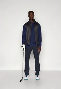 Golfo apranga, kurią sudaro juoda paminkštinta liemenė, tamsiai mėlyna ilgomis rankovėmis marškiniai, tamsios kelnės, balti golfo batai ir golfo lazda su balta pirštine.