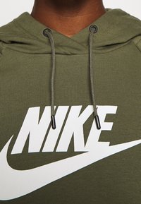 Olivgrön hoodie med en stor vit Nike-logotyp, dragsnören med svarta ändar och en texturerad tygfinish.