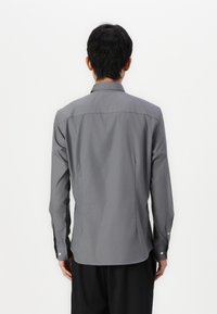 Camisa gris de manga larga con cuello estándar, puños con botón y dobladillo recto. Textura suave y corte entallado, mostrada desde la parte de atrás.