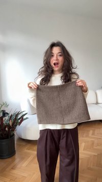 Swatch di tessuto a spina di pesce marrone in primo piano, con una superficie liscia. Sullo sfondo si intravede un divano curvo di colore chiaro e delle piante.