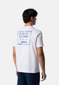 T-shirt di cotone bianco con maniche corte, caratterizzata da un design stampato blu sul retro che include motivi da spiaggia e testo blu.