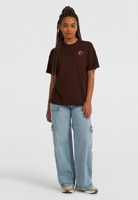 Hnedé bavlnené tričko s malou ružovou grafikou na ľavej strane hrude, v páre s svetlomodrými voľnými cargo džínsami s bočnými vreckami.