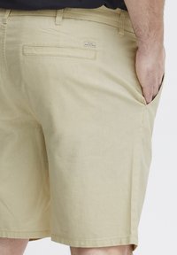 Lys beige shorts laget av et glatt, lett materiale. Har en baklomme og synlig sømdetalj langs kanten.