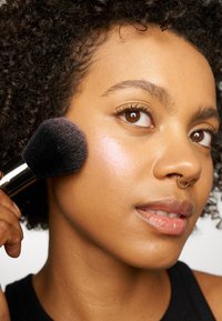 Pinceau à maquillage avec des poils noirs denses appliquant de l'enlumineur sur la joue. L'utilisateur a des cheveux bouclés, porte un haut noir et montre une texture de peau lisse.