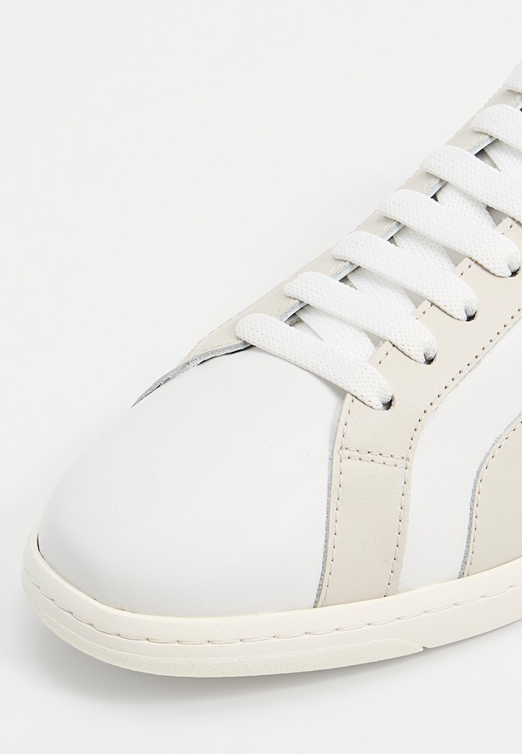 Białe skórzane sneakersy z beżowymi akcentami, białymi sznurówkami i gumową podeszwą, prezentowane z bliskiej perspektywy, skupiając się na czubku i obszarze sznurowania.
