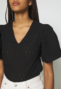 Černý vyšívaný top s výstřihem do V, krátkými puffovými rukávy, texturovanými květinovými vzory a otevřenými tkaninami, v kombinaci s světlými kalhotami.