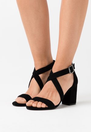 Sorte ruskind blockhælede sandaler med krydsende remme og ankelspænde. Open-toe design med en glat overflade.