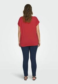 Červený top s krátkým rukávem a volným střihem, kombinovaný s tmavě modrými těsnými džíny a černými sandály. Materiál vypadá hladce a ležérně.