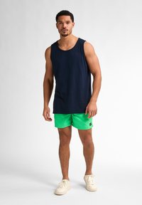 Top din bumbac de culoare bleumarin, asortat cu șorturi verzi strălucitoare. Modelul poartă adidași crem, evidențiind o ținută casual de vară.