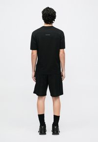 Fekete rövid ujjú ing texturált hátrésszel, fekete rövidnadrággal párosítva, amely finom hatszögletű mintázattal rendelkezik. Feketén sportcipővel és zoknival viselve.