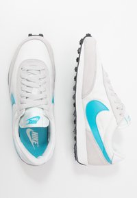 Bílé a šedé tenisky s modrým swoosh, s tkaničkovým designem, gumovou podrážkou a kombinací semišových a textilních materiálů.