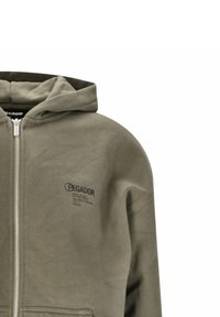 Felpa con zip color verde oliva, dotata di tasche frontali, polsini a costine e cappuccio. Presenta il logo sul lato sinistro del petto. Il materiale sembra morbido e con una trama interessante.