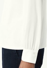 Weißes Langarmshirt mit gerafften Ärmeln; weicher Baumwollstoff; ausgestattet mit einer knopfbefestigten Manschette; kombiniert mit dunklen Jeanshosen.