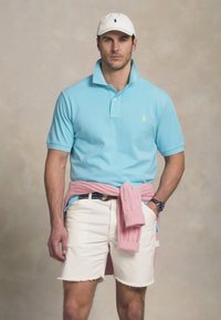 Ανοιχτό μπλε κοντομάνικο πουκάμισο polo με μικρό λογότυπο, άσπρα τζιν σορτς με φθαρμένες άκρες, και μια ροζ ζακέτα δεμένη στη μέση.