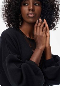 Czarny, oversize sweter z bufiastymi rękawami, wykonany z teksturowanej tkaniny, nałożony na przezroczystą bluzkę z kwiatowymi wzorami. Modelka delikatnie zaciska dłonie.