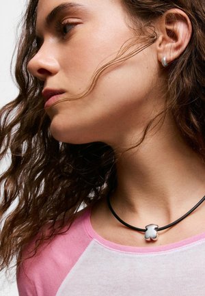 Joven con cabello rizado y castaño que lleva un colgante plateado con forma de oso en un collar de cordón negro y una camiseta rosa y blanca.