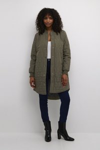 Lång olivgrön vadderad jacka med rund hals, tryckknappar och ribbade ärmslut. Kombinerad med mörka jeans och svarta klackstövlar.