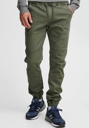 Olive green jogger pants with an elastic waistband, drawstring, cuffed ankles, and knee panel details, paired with navy athletic shoes.