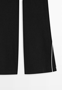 Zwarte broek met rechte brede pijpen en een witte verticale streep langs de buitennaad van het rechterpijp.