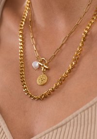 Goldfarbene mehrschichtige Halsketten mit einer strukturierten Kette, einem Anhänger mit einem Augenmotiv und einem Perlenakzent.