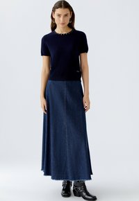 Navy gestricktes Oberteil mit kurzen Ärmeln und dekorativen Perlen am Ausschnitt, kombiniert mit einem dunkelblauen Denim-Maxirock mit rohem Saum und Vordernaht. Schwarze Schnallenstiefel.