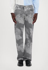 Jean en denim gris avec un motif répété de visages de chérubins et d'ailes, porté avec des chaussures noires et une chemise à manches longues bleu clair.