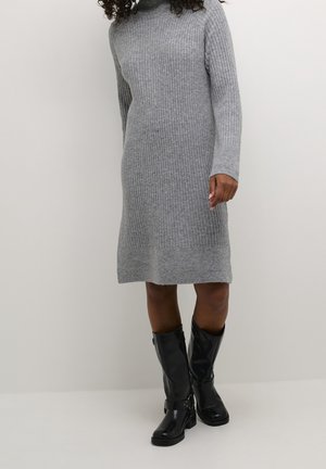 Femme portant une robe longue à manches longues en tricot côtelé gris, associée à des bottes en cuir noires montant jusqu'aux genoux, debout devant un fond uni.