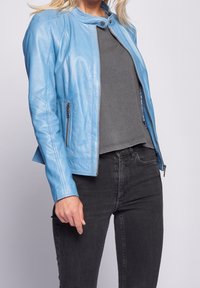 Kvinde iført en lys blå læderjakke over en grå top, kombineret med sorte jeans, stående mod en ensfarvet lys baggrund.