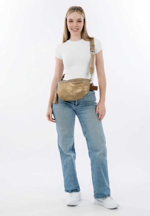 Sac banane en or, métallique, avec une fermeture zippée, un détail de gland et une sangle ajustable, porté à la taille avec une tenue décontractée.