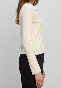 Žebrovaný krémový cardigan s límečkem a zapínáním na knoflíky. Má dlouhé rukávy a boční rozparky, vyrobený z jemného pleteného materiálu.