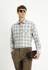 Męska koszula w kratę z długim rękawem w odcieniach bieli, niebieskiego i brązowego, z kieszonką na piersi, w połączeniu z brązowymi spodniami z sztruksu i brązową torbą.