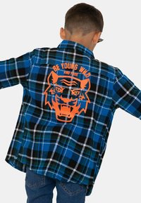 Blau-schwarz kariertes Flanellhemd mit einem orangenen Tiger-Graphic und dem Text "SEI JUNG, WILD UND FREI" auf der Rückseite.