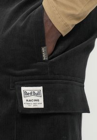 Fekete kordbársony overálok, elülső zsebbel, texturált anyaggal és egy fehér címkével, amelyen a "Red Bull Racing" felirat és a "2004" évszám szerepel.