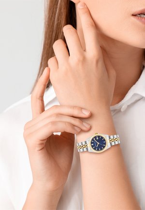 Montre métallique argent et or avec un cadran bleu, doté d'une lunette texturée, d'un bracelet en maille et d'une fonction de date, portée sur un poignet.