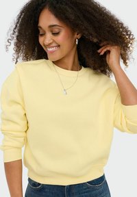 Femme aux cheveux bouclés portant un sweat-shirt jaune pâle et un collier argenté, souriante et touchant ses cheveux, sur un fond uni.